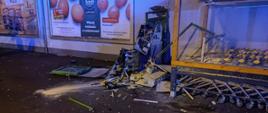 Fotografia przedstawia kompletnie zniszczony wybuchem bankomat znajdujący się przy markecie Biedronka, obok znajdują się wózki sklepowe. Na tylnej ścianie budynku widać banery reklamowe.