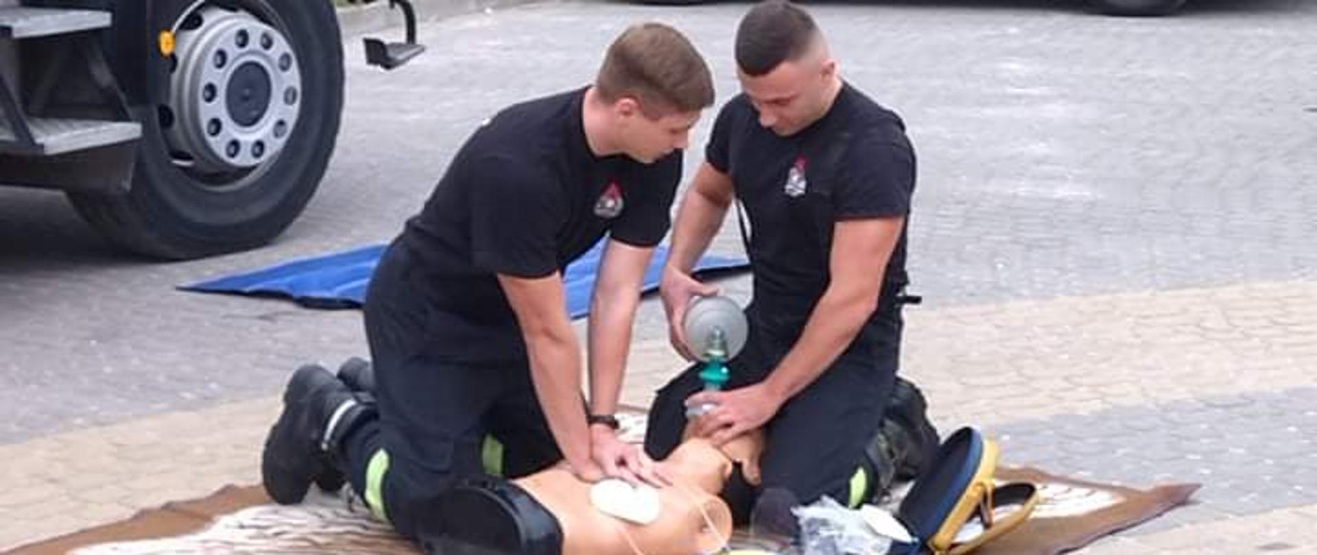 Zdjęcie obrazuje strażaków pokazujących w jaki sposób należy udzielać kwalifikowanej pierwszej pomocy przy użyciu sprzętu ratownictwa medycznego. Pokaz odbywa się przy pomocy fantoma. Obok nich stoi samochód pożarniczy , a za nimi samochód osobowy i w tle widać roślinność.