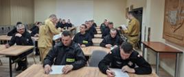Strażacy ubrani w czarne ubrania koszarowe w trakcie egzaminu teoretycznego w Sali szkoleniowej, siedzą przy ławkach na których leżą białe karty egzaminacyjne, ponadto w tle widać biały ekran rzutnika 