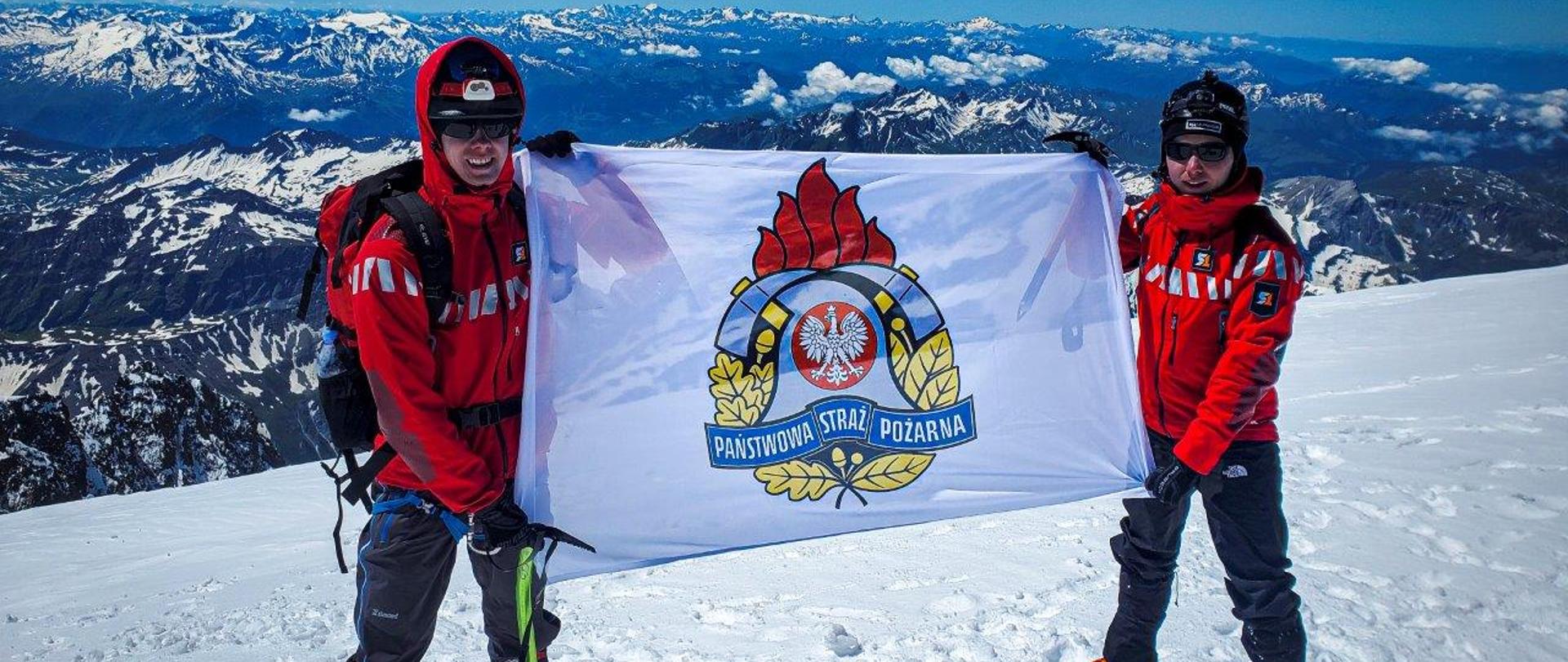 Strażacy zdobyli Mont Blanc dla Fundacji Dorastaj z Nami – na zdjęciu dwóch strażaków w alpinistycznych ubraniach z flagą z godłem PSP – w tle rozległa panorama górska.