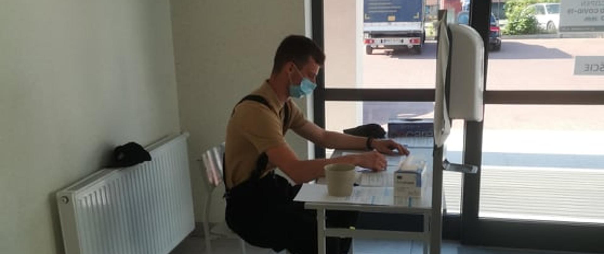 Punkt szczepień w Rawiczu. Przy stoliku siedzi strażak, który przegląda dokumentacje. Obok stolika stoi dozownik środka dezynfekcyjnego. Przez przeszklone drzwi widać pobliski blok mieszkalny.