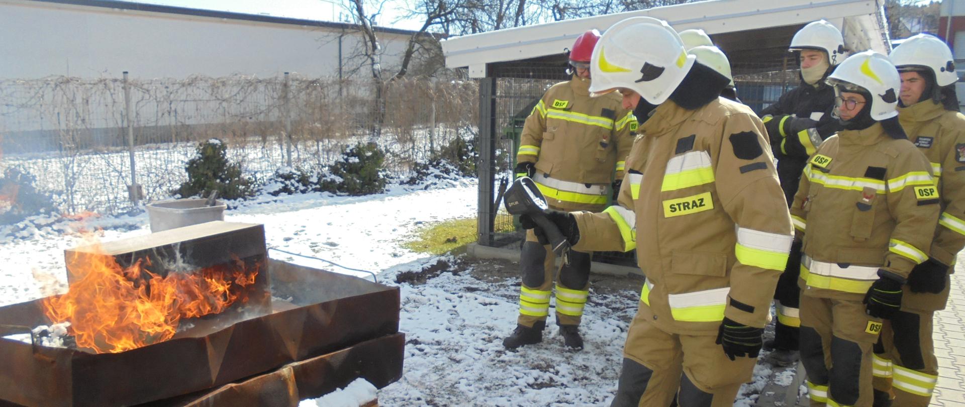 Na zdjęciu widać strażaka w trakcie ćwiczeń związanych z tematyką pożarów budynków mieszkalnych. Jest pora dzienna. Ma zdjęciu widać siedmiu strażaków którzy mają założone ubranie specjalne- spodnie i kurtkę, chroniących ich m. in. przed działaniem wysokiej temperatury oraz czerwony i białe hełm strażacki na głowie. Po lewej stronie widać palący się domek drewniany z którego otworów wychodzi płomień i widać dym. Domek ustawiony jest na platformie z palet drewnianych i stoi na wannach. Jeden strażak trzyma w ręce kamerę termowizyjną skierowany w stronę ognia. Ćwiczenia odbywają się w plenerze na trawie.