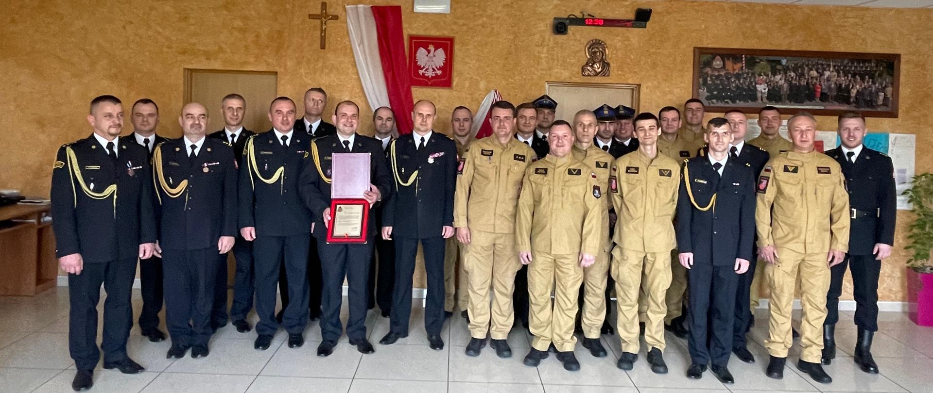 Grupa 25 funkcjonariuszy Państwowej Straży Pożarnej pozuje do wspólnego zdjęcia z okazji przejścia na zaopatrzenie emerytalne jednego z funkcjonariuszy. Zdjęcie zrobione w pomieszczeniu o żółto-brązowych ścianach w jednostce ratowniczo-gaśniczej nr 2 w Mielcu. Na środku stoją strażacy ubrani w galowe mundury w kolorze czarnym oraz mundury koszarowe w kolorze piaskowym. Niektórzy mają przypięte odznaczenia, a kilku strażaków nosi czapki służbowe.
Na pierwszym planie, w środku grupy, mężczyzna w czarnym mundurze trzyma pamiątkową tablicę oraz teczkę – dokument potwierdzający przejście na emeryturę. Wszyscy stoją w pozycji wyprostowanej, z rękami ułożonymi wzdłuż ciała lub za plecami, patrząc w stronę aparatu.
W tle widoczne są biało-czerwona flaga Polski, godło państwowe oraz krzyż wiszący na ścianie. Nad drzwiami wisi zegar elektroniczny wskazujący godzinę 12:39. Po prawej stronie znajduje się duże, oprawione zdjęcie grupowe oraz rośliny doniczkowe ustawione przy ścianie.
Zdjęcie zostało wykonane w jasnym, dobrze oświetlonym pomieszczeniu o białej, kafelkowej podłodze.
