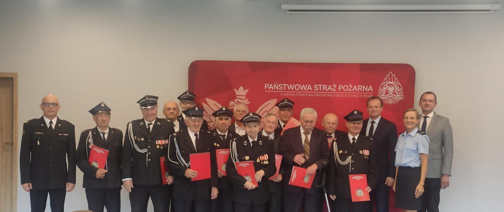 Zdjecie grupowe na tle baneru PSP Śrem na którym są goście oraz druhowie OSP