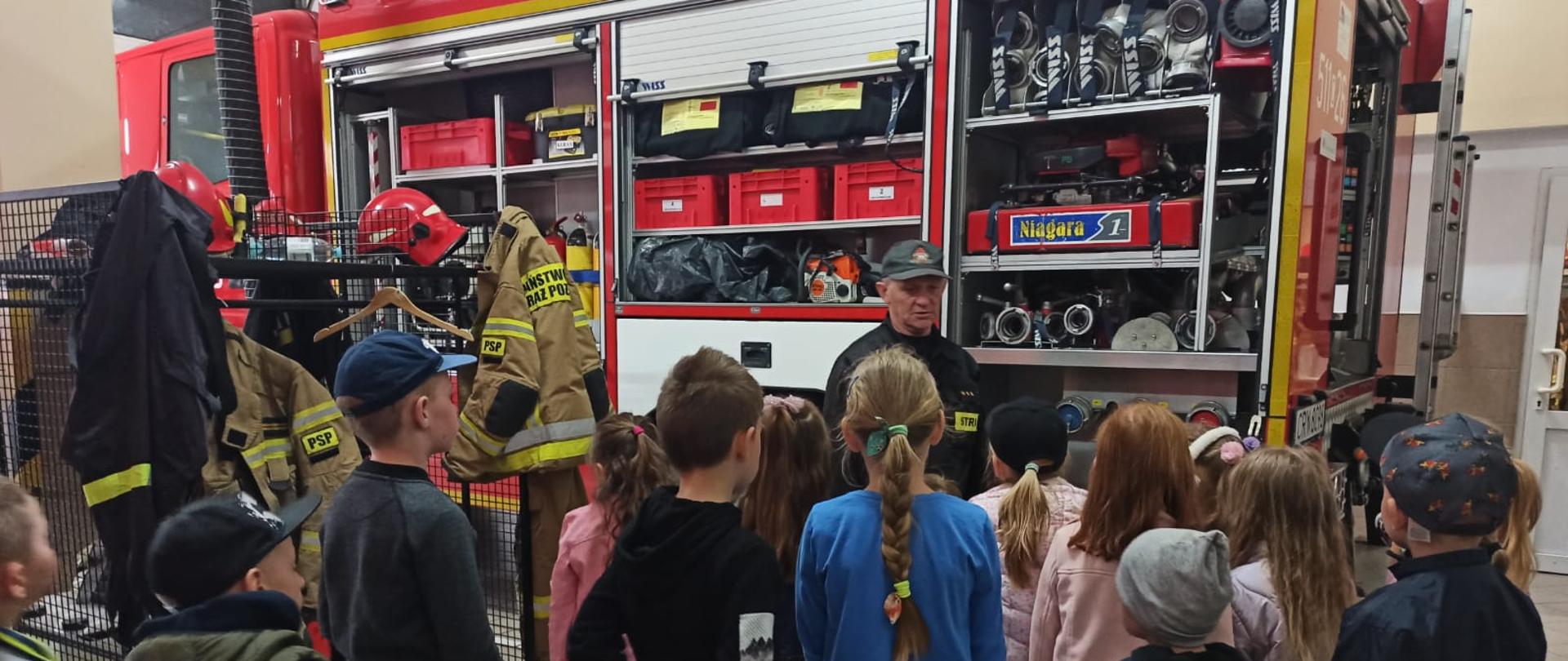 Zdjęcie przedstawia odwrócone tyłem dzieci, które oglądają sprzęt w w samochodzie pożarniczym, który znajduje się w garażu. 