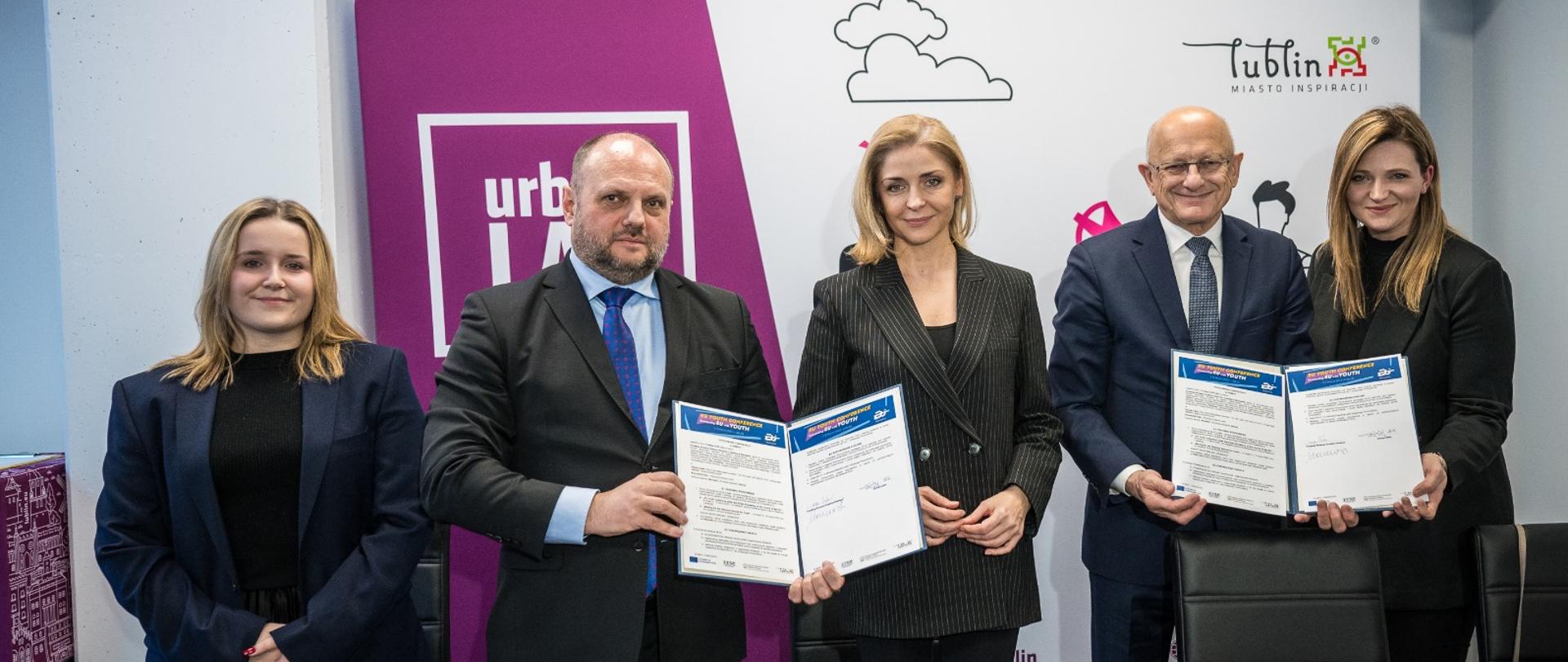 Pięć osób stoi i pozuje do zdjęcia. Trzecią osobą jest wiceminister Joanna Mucha. W rękach trzymają teczki z podpisanym porozumieniem. 