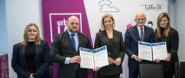 Pięć osób stoi i pozuje do zdjęcia. Trzecią osobą jest wiceminister Joanna Mucha. W rękach trzymają teczki z podpisanym porozumieniem. 