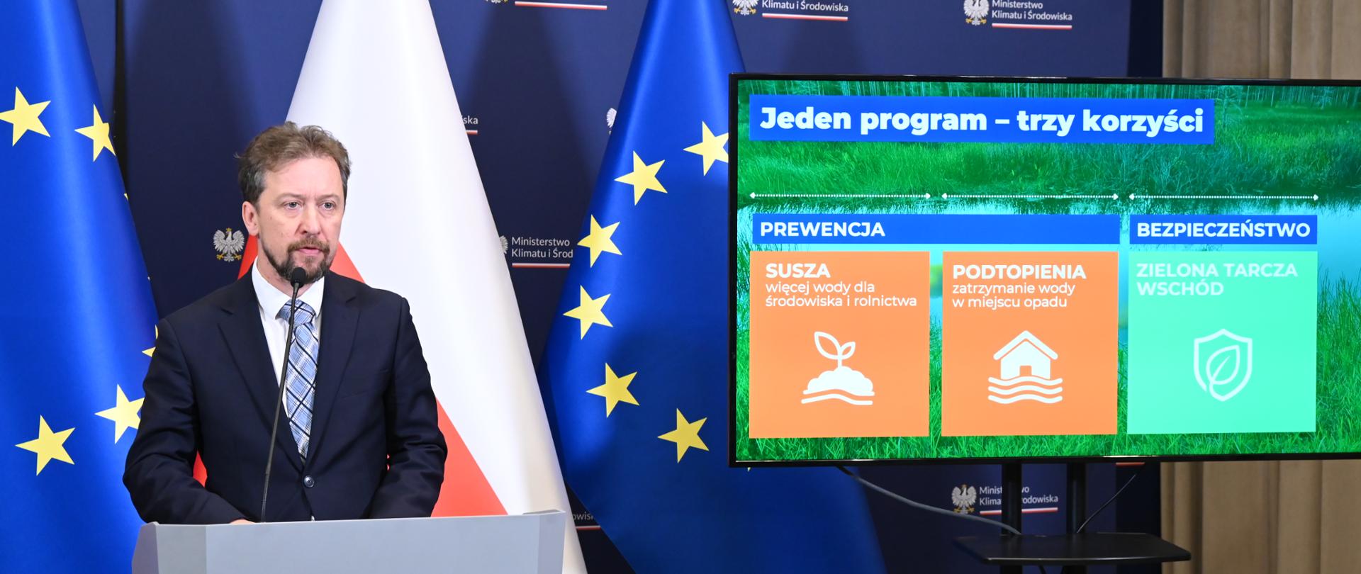 Zdjęcie przedstawia wystąpienie podczas konferencji prasowej poświęconej ochronie środowiska. Przy mównicy widoczny jest Piotr Otawski, Generalny Dyrektor Ochrony Środowiska. W tle znajduje się flaga Polski i Unii Europejskiej oraz ścianka Ministerstwa Klimatu i Środowiska. Na ekranie obok wyświetlono slajd prezentujący korzyści programu związane z przeciwdziałaniem suszy, ograniczaniem podtopień oraz wzmacnianiem bezpieczeństwa środowiskowego.