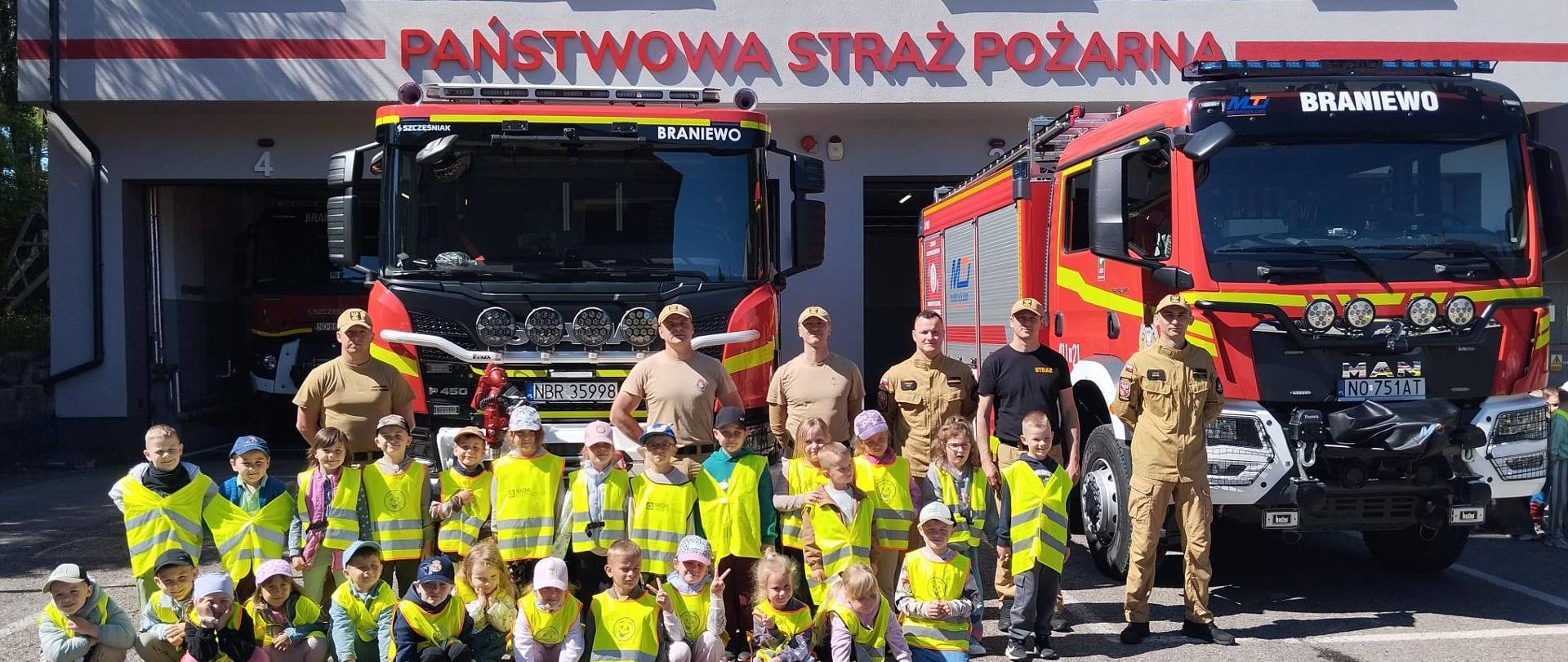 Grupa przedszkolaków w żółtych kamizelkach pozuje do zdjęcia ze strażakami przy dwóch dużych samochodach strażackich. Słonecznie.