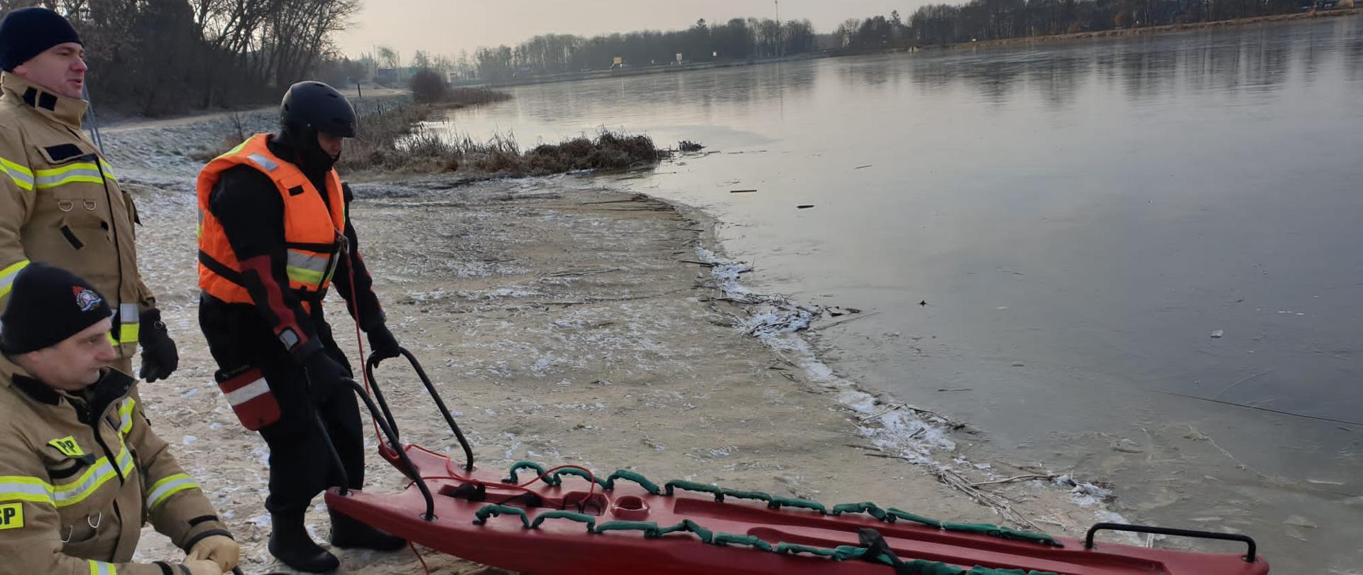 Strażacy stoją na plaży i przygotowują się do ćwiczeń. Dwóch jest ubranych w ubrania specjalne, jeden z nich trzyma linę na kołowrotku przymocowaną do sań lodowanych trzymanych przez strażaka ubranego w suchym skafandrze.