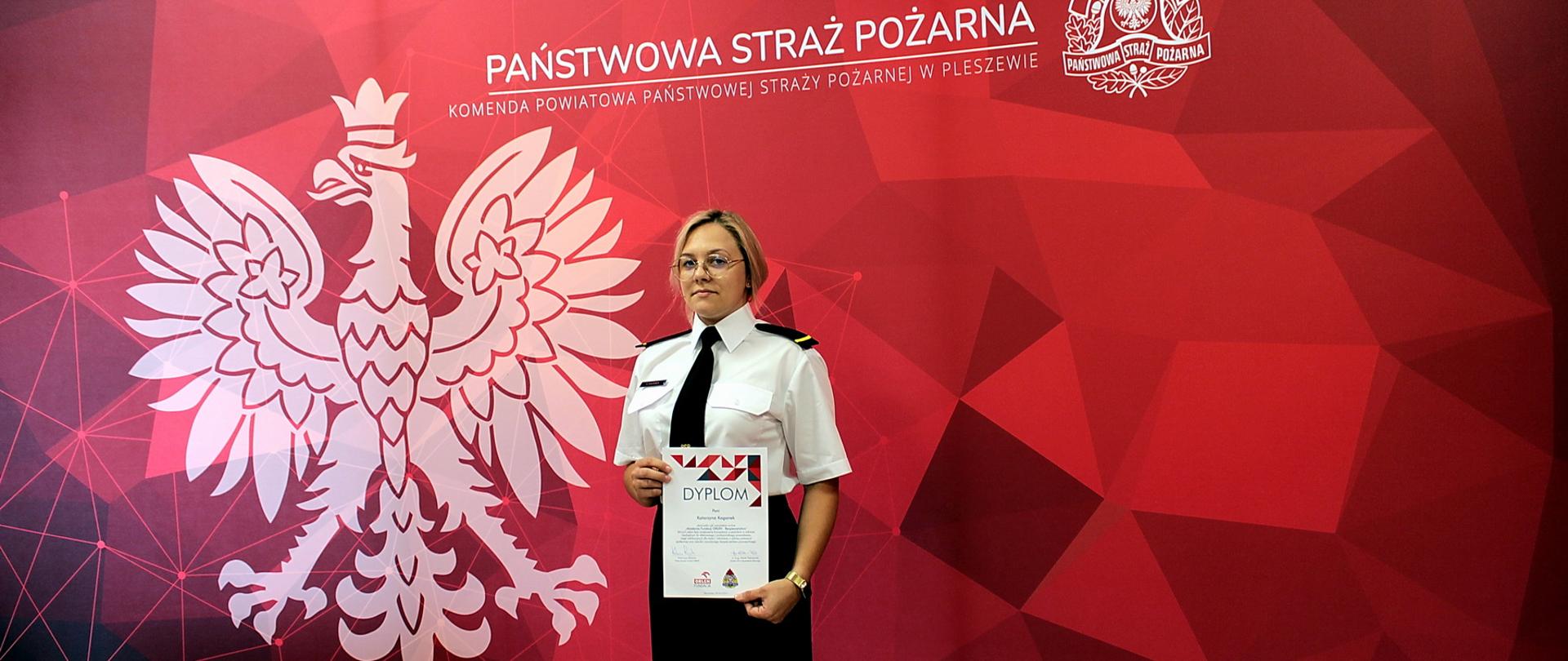 Kobieta w mundurze wyjściwoym PSP stoi przed banerem straży pożarnej, na którym widoczny jest orzeł państwowy oraz logo PSP i napis Państwowa Straż Pożarna na czerwonym tle. Kobieta trzyma w ręku dyplom