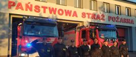 Ośmiu strażaków ustawionych w szeregu na tle dwóch pojazdów pożarniczych przed budynkiem komendy oddają hołd zmarłemu strażakowi śp. ogn. Pawłowi Chyle z KM PSP w Grudziądzu. Dowódca salutuje. Pojazdy posiadają włączone niebieskie światła błyskowe.