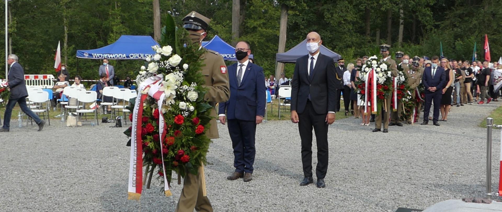 Na pierwszym planie żołnierz trzymający wieniec od prezydenta RP, za nim stoją wojewoda opolski Adrian Czubak i dyrektor biura wojewody Mariusz Nieckarz, z tyłu rząd ludzi ustawionych, oczekujących na złożenie kwiatów. W tle widać drzewa.