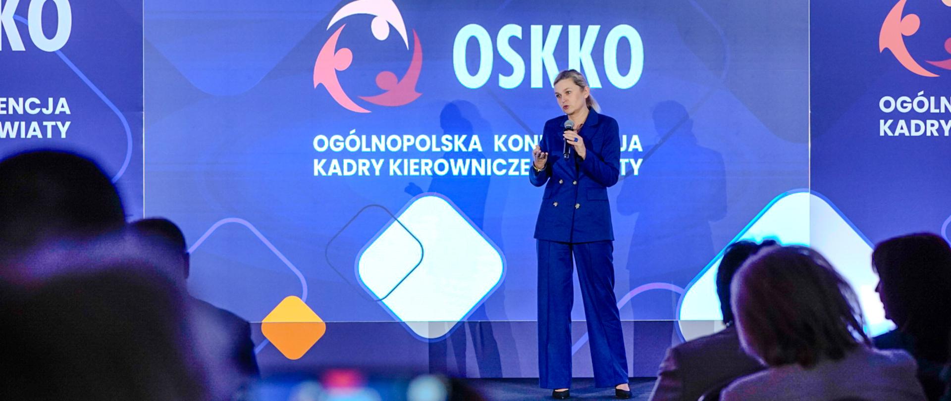 Minister Edukacji Barbara Nowacka przemawia na scenie podczas XXI Konferencji OSKKO. Ma na sobie niebieski garnitur, trzyma mikrofon i gestykuluje. W tle widoczne logo OSKKO.