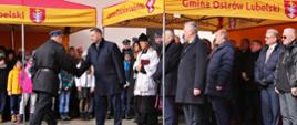 Na podwórku wyłożonym kostka stoją żółto-czerwone daszki namiotowe z napisem Gmina Ostrów Lubelski, pod nimi duża grupa ludzi, na środku minister Czarnek podaje rękę mężczyźnie w czarnym mundurze.