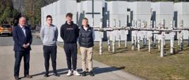 Trzej uczniowie i nauczyciel stojący na cmentarzu, w tle widoczny duży pomnik i liczne brzozowe krzyże.