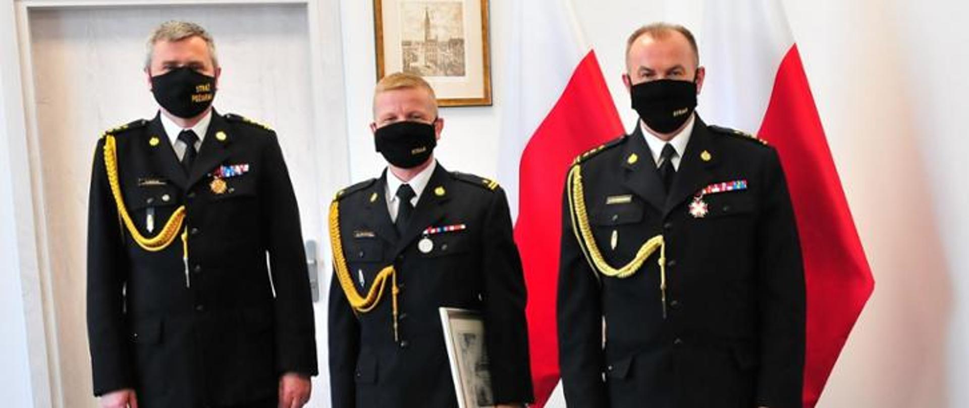 na zdjęciu widoczni są 3 funkcjonariusze PSP w umundurowaniu wyjściowym w tle dwie flagi biało-czerwone i wiszący obrazek na ścianie