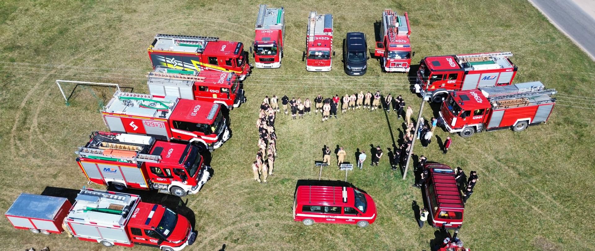 Zdjęcie zrobione z lotu ptaka. Na zdjęciu widać samochody pożarnicze, które ustawione są w kształcie litery U. Wewnątrz pojazdów strażacy stoją w szyku zwartym podczas odprawy.