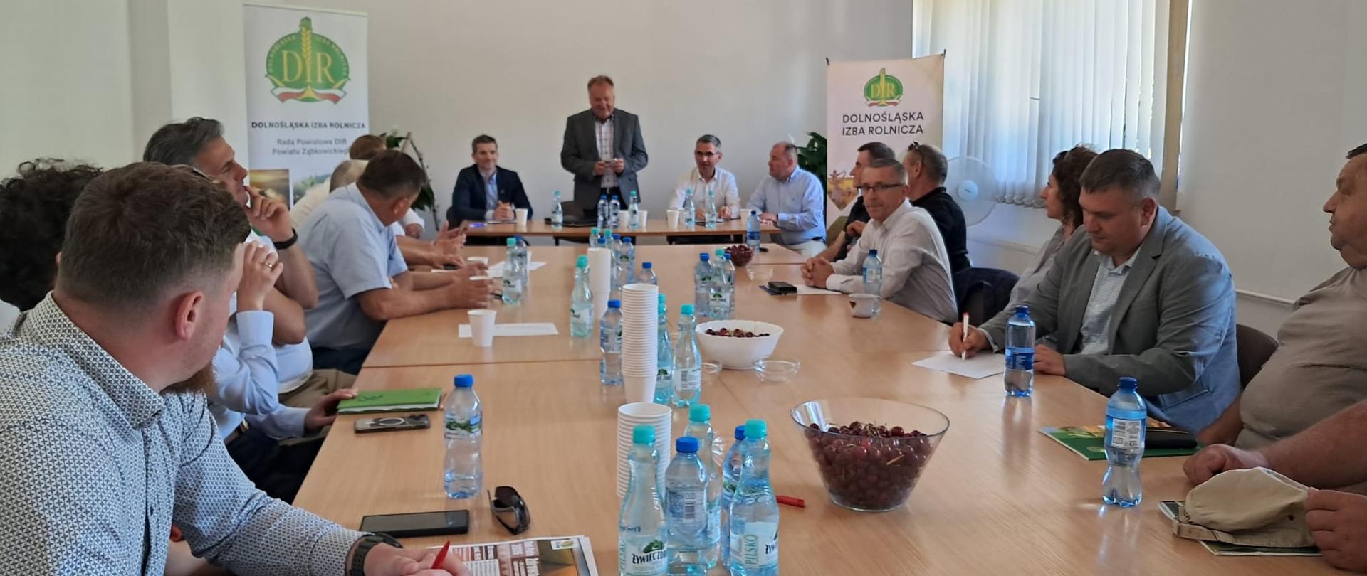 Zdjęcie przedstawia spotkanie w jasnej sali konferencyjnej z wysokim sufitem i dużymi oknami po prawej stronie. Wokół długiego drewnianego stołu siedzi kilkanaście osób, większość notuje lub słucha prowadzącego. Na końcu stołu stoi mężczyzna w marynarce, który przemawia do zgromadzonych. Na stole znajdują się butelki z wodą, dokumenty, notatniki, długopisy oraz miski z owocami. Po obu stronach prowadzącego ustawiono banery z logo i nazwą Dolnośląskiej Izby Rolniczej. 