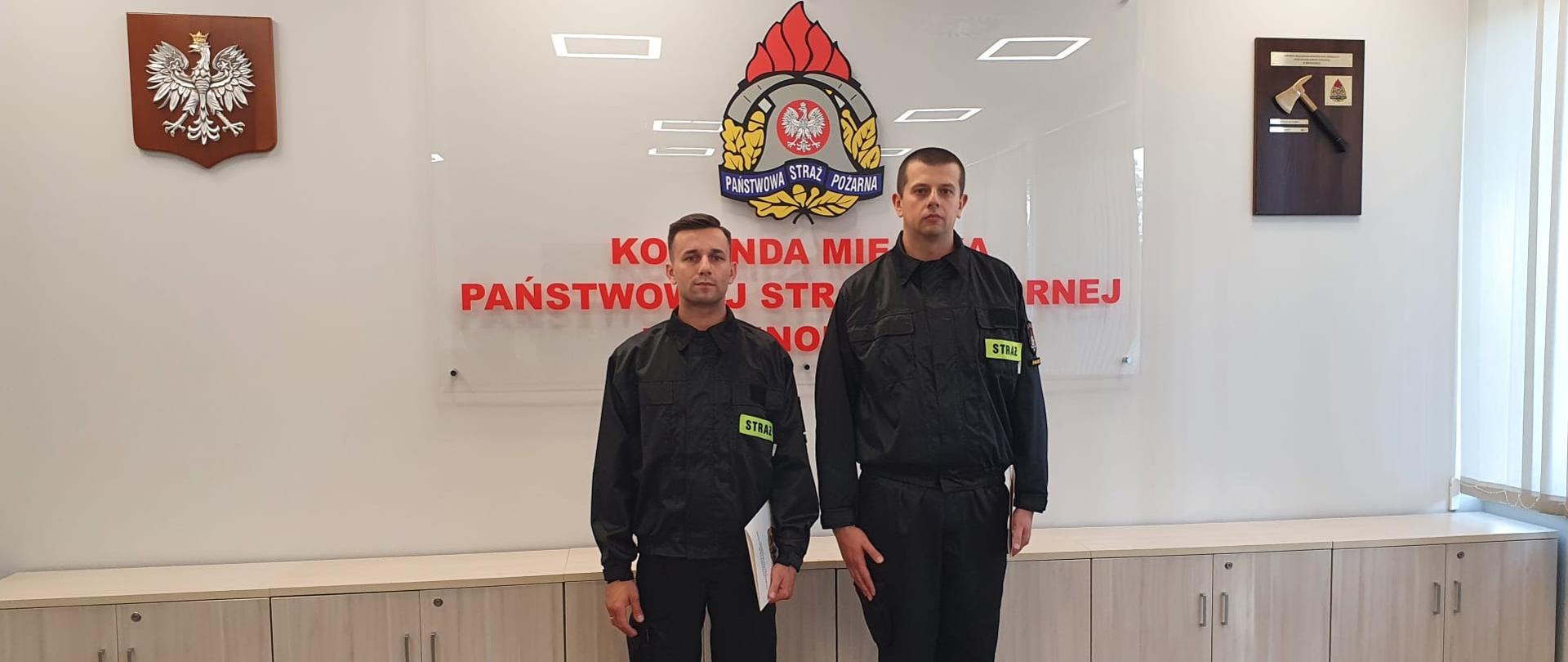 Na zdjęciu widoczni funkcjonariusze w sali konferencyjnej w komendzie miejskiej Państwowej Straży Pożarnej w Świnoujściu trzymający w rękach akty ślubowania. Stoją na tle godła, logo psp oraz napisu Komenda Miejska Państwowej Straży Pożarnej w Świnoujściu.