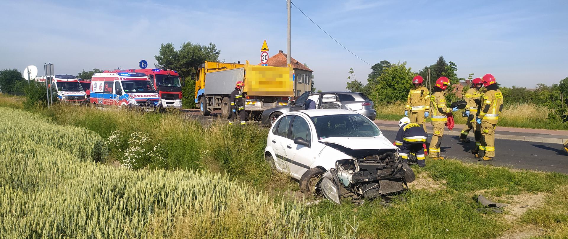 Na pierwszym planie biały samochód marki citroen z uszkodzonym przodem. Obok pojazdu, który znajduje się na poboczu drogi stoją strażacy w ubraniach bojowych. W tle widać rozbity drugi samochód oraz ciężarówkę. Po lewej stronie na pasie drogi stoją dwie karetki pogotowia oraz pojazd gaśniczy. 