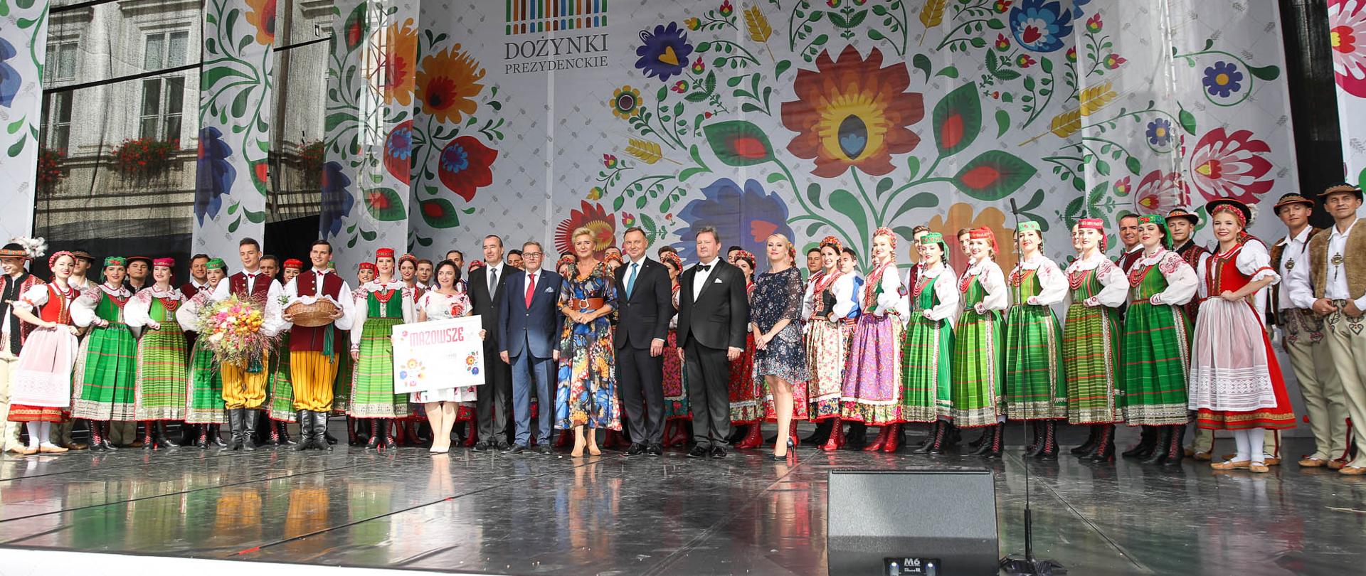 Scena, na której stoi duża grupa ludzi w strojach ludowych, w środku Prezydent RP z małżonką. 