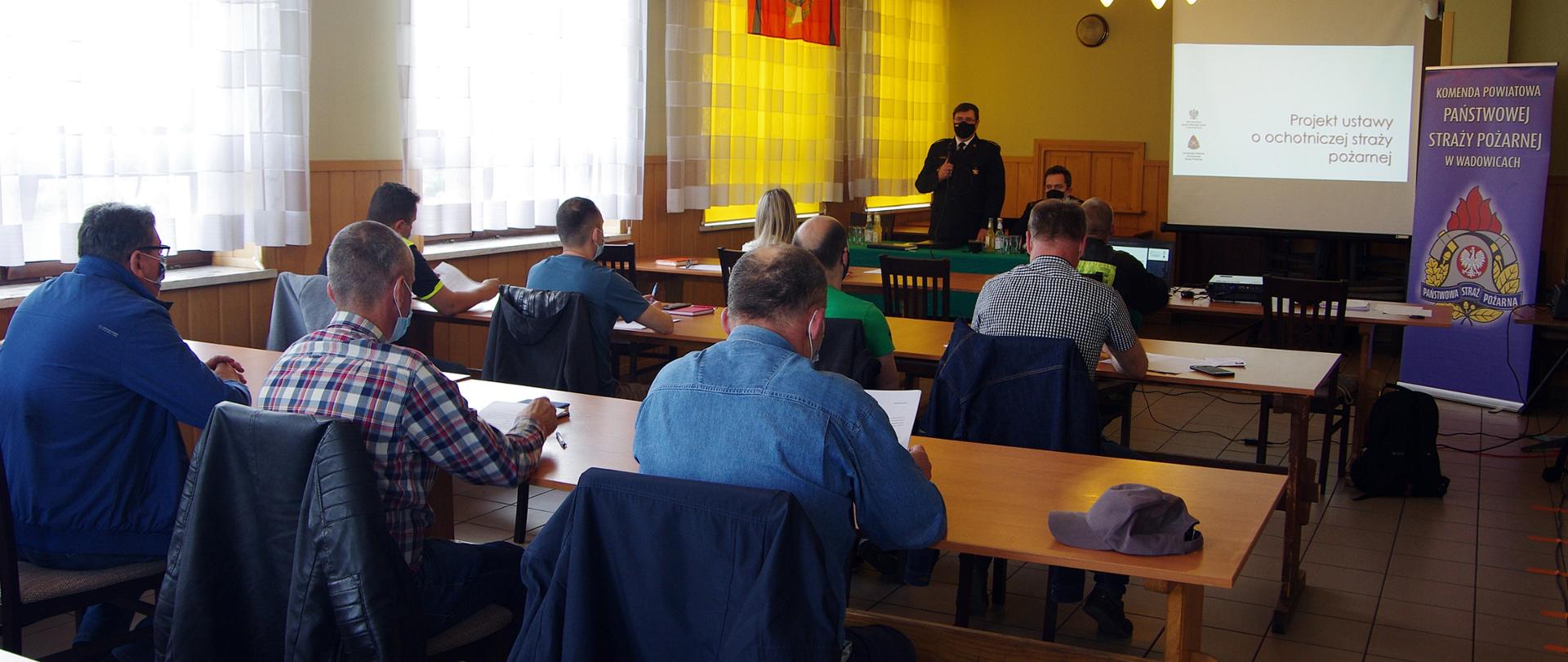Widok z głębi sali. Na pierwszym planie, za stołami na sali OSP, siedzą uczestnicy spotkania w maseczkach ochronnych. W tle widoczny stół prezydialny, za którym przodem do uczestników stoi w czarnym mundurze wyjściowym i maseczce ochronnej przemawiający komendant powiatowy PSP. Obok za stołem siedzi w czarnym mundurze wyjściowym i maseczce ochronnej zastępca komendanta powiatowego PSP. Po prawej stronie ekran z tekstem projektu ustawy o PSP oraz baner KP PSP w Wadowicach. 