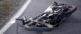 Zderzenie auta osobowego z motorowerem. Droga powiatowa. Na jezdni leży motorower uczestniczący w wypadku. Wokół niego plamy płynów eksploatacyjnych. 