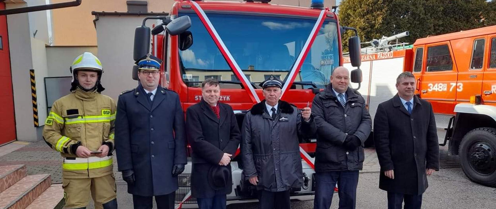 Na zdjęciu widać pojazd ratowniczo-gaśniczy na podwoziu Man z zabudową firmy Stolarczyk. Pojazd czerwony. Posiada białe oznaczenia. Posiada sygnalizację ostrzegawczą. Żaluzje zamknięte na zabudowie. W tle budynek OSP Rajsko. Pojazd stoi na asfalcie. Pora dnia to dzień. Przed pojazdem stoją osoby, które dokonywały przekazania pojazdu.