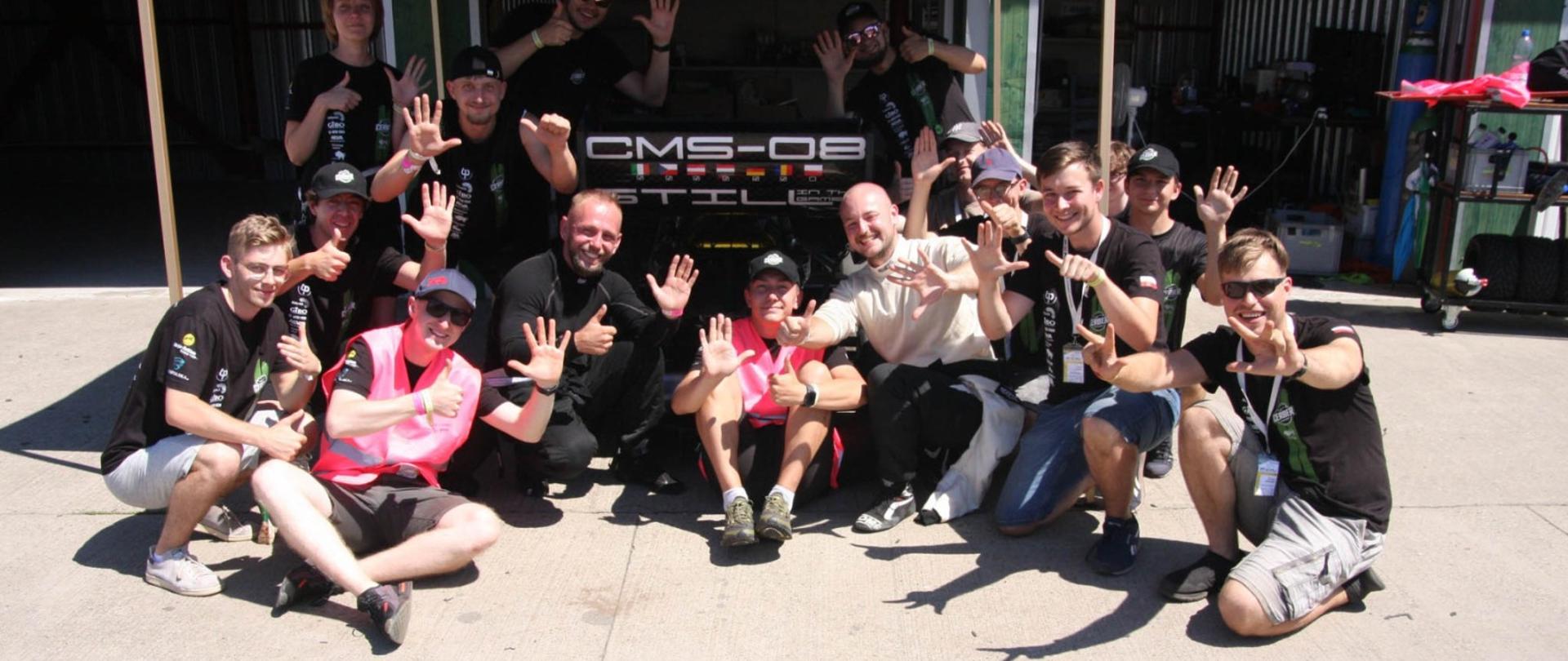 W ostrym słońcu na torze siedzi kilkanaście osób, za nimi pod daszkiem bolid CMS-08.