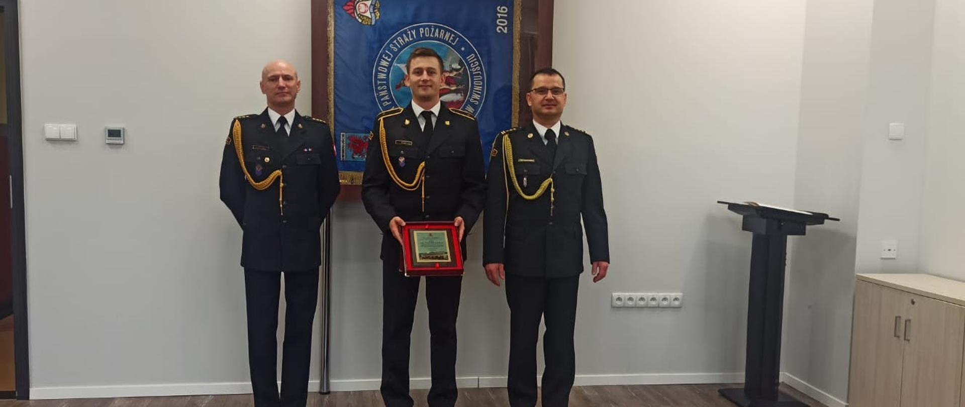Zdjęcie przedstawia 3 funkcjonariuszy KM PSP w Świnoujściu umundurowanych w mundury wyjściowe z sznurami. Od lewej Komendant Miejski st. kpt. Marek Wieczorek, dowódca sekcji asp. Paweł Łoś oraz Zastępca Komendanta Miejskiego st. bryg. Tomasz Wojciechowski. Strażacy stoją przy ścianie koloru białego na tle sztandaru Komendy Miejskiej Państwowej Straży Pożarnej w Świnoujściu