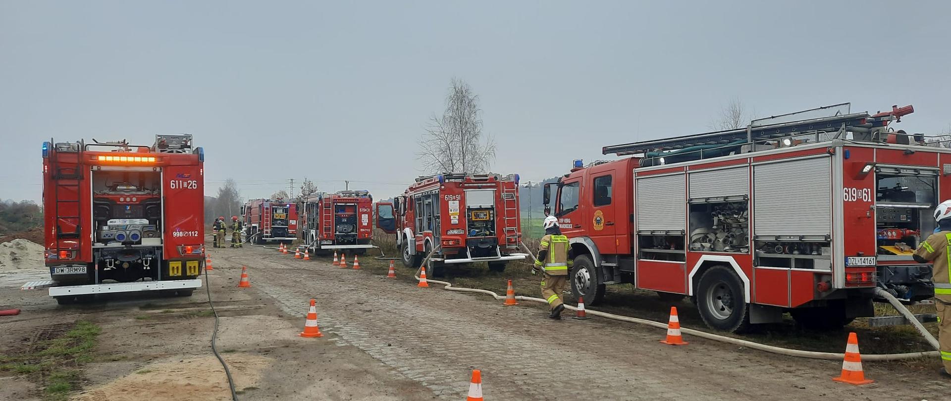 widoczne pojazdy gaśnicze stojące jeden za drugim z rozwiniętymi wężami, w ich otoczeniu umundurowani strażacy