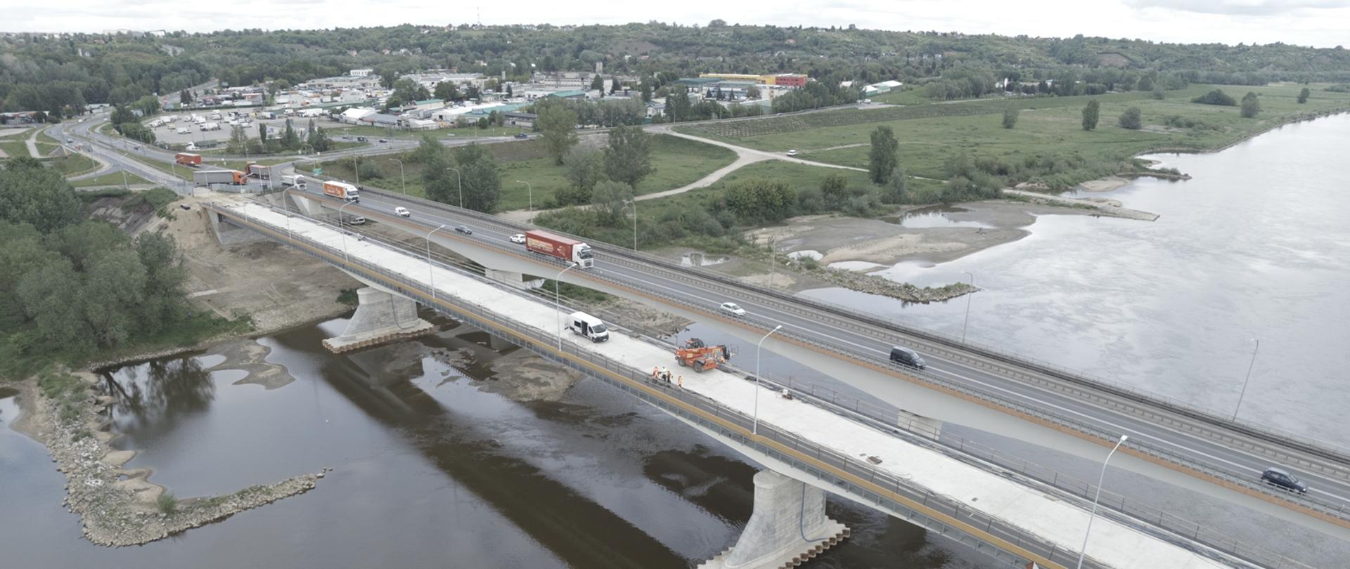 Most w Sandomierzu - dwa mosty nad rzeką, zdjęcie z drona. Po jednym jeżdżą samochody, drugi równoległy w budowie. Na budowanym moście biały beton, maszyna budowlana, pracownicy. W oddali zabudowania miejskie 