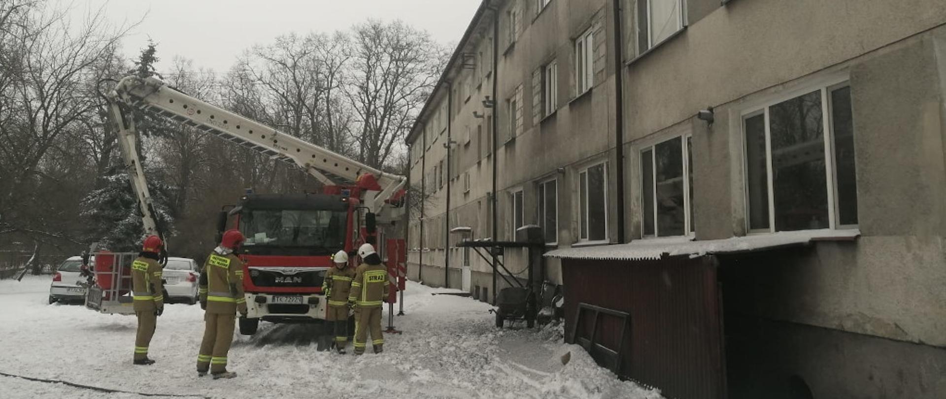 Na zdjęciu przedstawiony jest przód specjalnego samochodu pożarniczego z podnośnikiem hydraulicznym. Pojazd rozłożone ma podpory hydrauliczne oraz przygotowany park drabinowy do wjazdu na dach budynku. Przed samochodem do działań gaśniczych przygotowują się czterej strażacy w umundurowaniu specjalnym. Dwóch z nich na głowie ma założone hełmy koloru czerwonego, natomiast druga dwójka- koloru białego. Po prawej stronie znajduje się frontowa część budynku domu dziecka.