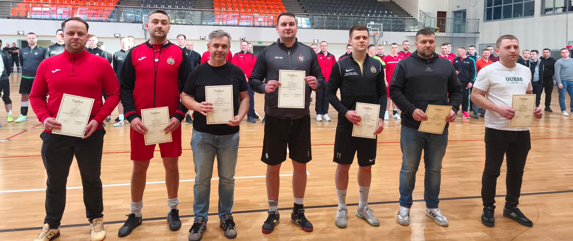 Na zdjęciu widać grupę strażaków reprezentujących różne jednostki, którzy pozują z pamiątkowymi dyplomami na parkiecie hali sportowej. Wydarzenie odbywa się w profesjonalnym obiekcie z trybunami, co podkreśla oficjalną rangę eliminacji strefowych w futsalu.