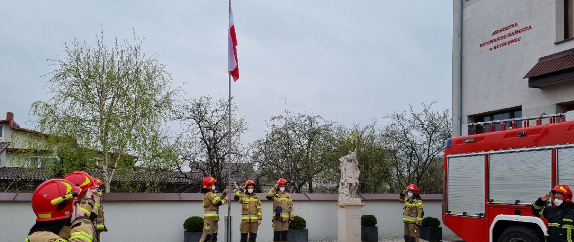 Uroczyste podniesienie flagi w Komendzie Powiatowej PSP w Szydłowcu - strażacy wciągający flagę państwową na maszt oddający honor fladze przez salutowanie.