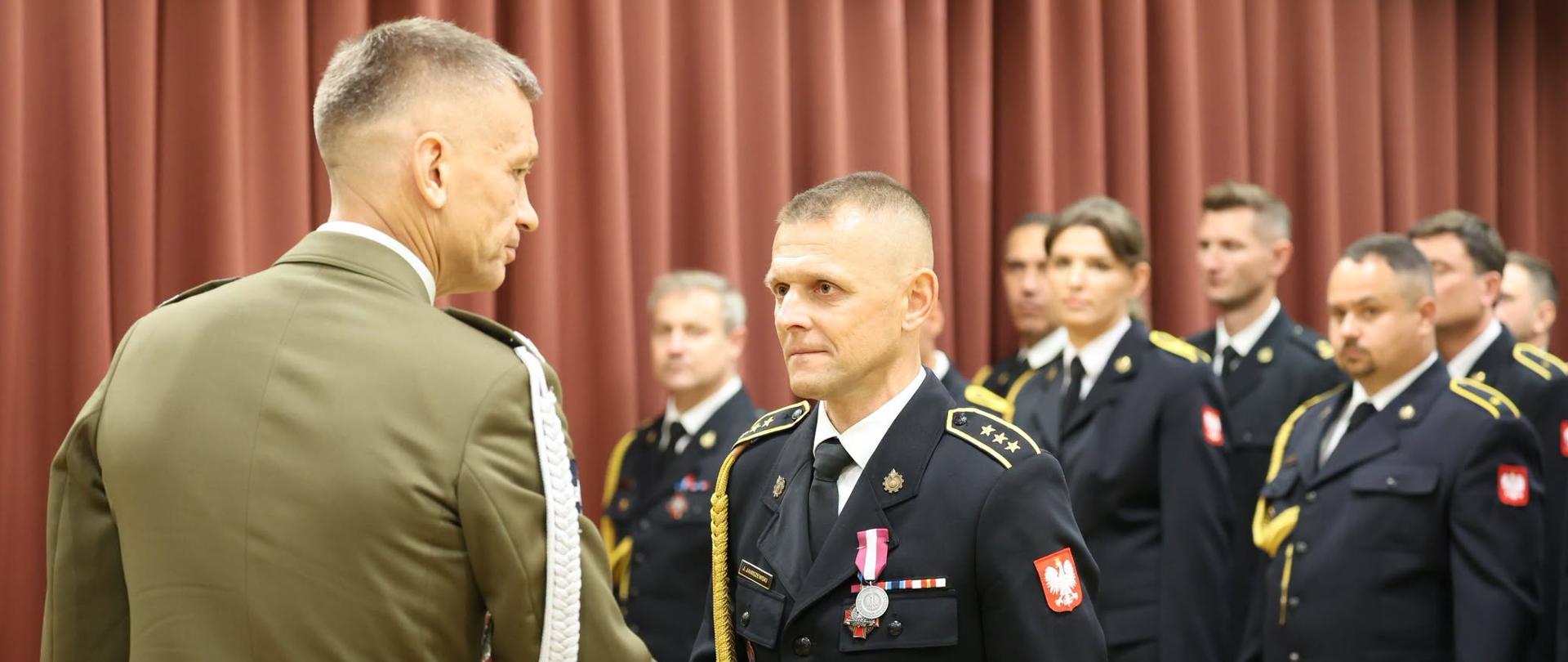 Wręczenie medalu odznaczonemu funkcjonariuszowi KP PSP w Makowie Mazowieckim przez Dyrektora Departamentu Zwierzchnictwa nad Siłami Zbrojnymi i Służb Mundurowych Biura Bezpieczeństwa Narodowego. W oddali widać pododdział odznaczonych. Wszyscy są ubrani w umundurowanie wyjściowe. 