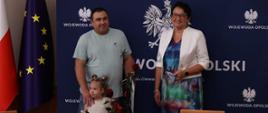 Wojewoda Opolski Monika Jurek razem z nowymi obywatelami polskimi pozują do zdjęcia. Jest to dorosły mężczyzna oraz kilkuletnia dziewczynka, jego córka. Oboje trzymają w dłoniach róże. 