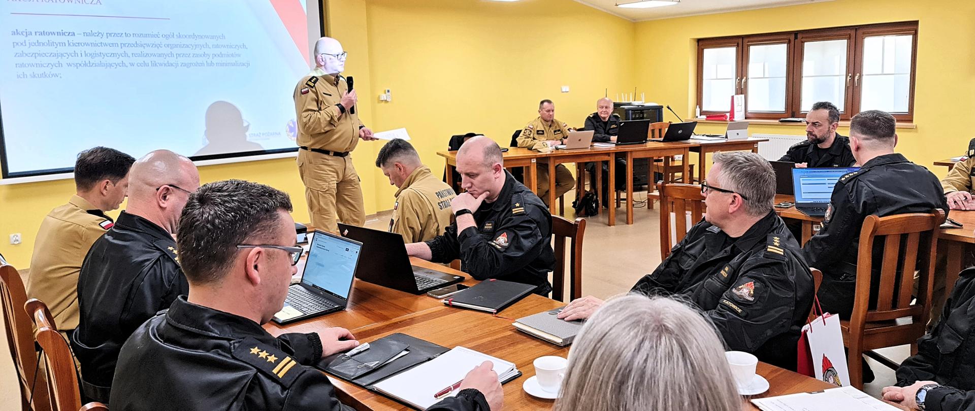 Sala konferencyjna z uczestnikami i prowadzącym szkolenie dyrektorem Biura Edukacji. W kadrze centralnym widoczne są trzy rzędy stołów, przy których siedzą uczestnicy narady. 