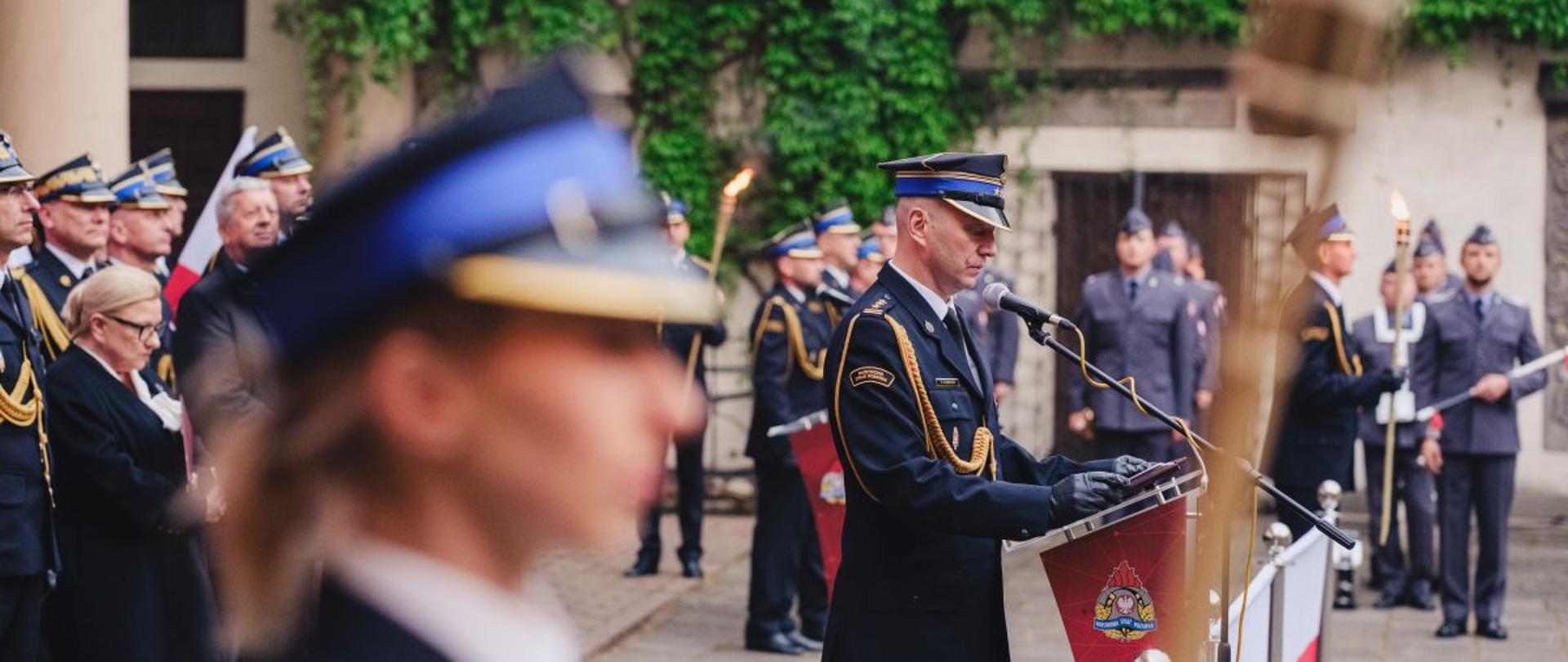 Funkcjonariusz PSP w galowym mundurze stoi przy podium z emblematem straży pożarnej i wygłasza przemówienie. Za nim stoją strażacy z pochodniami, a tło stanowi kamienna ściana porośnięta bluszczem.