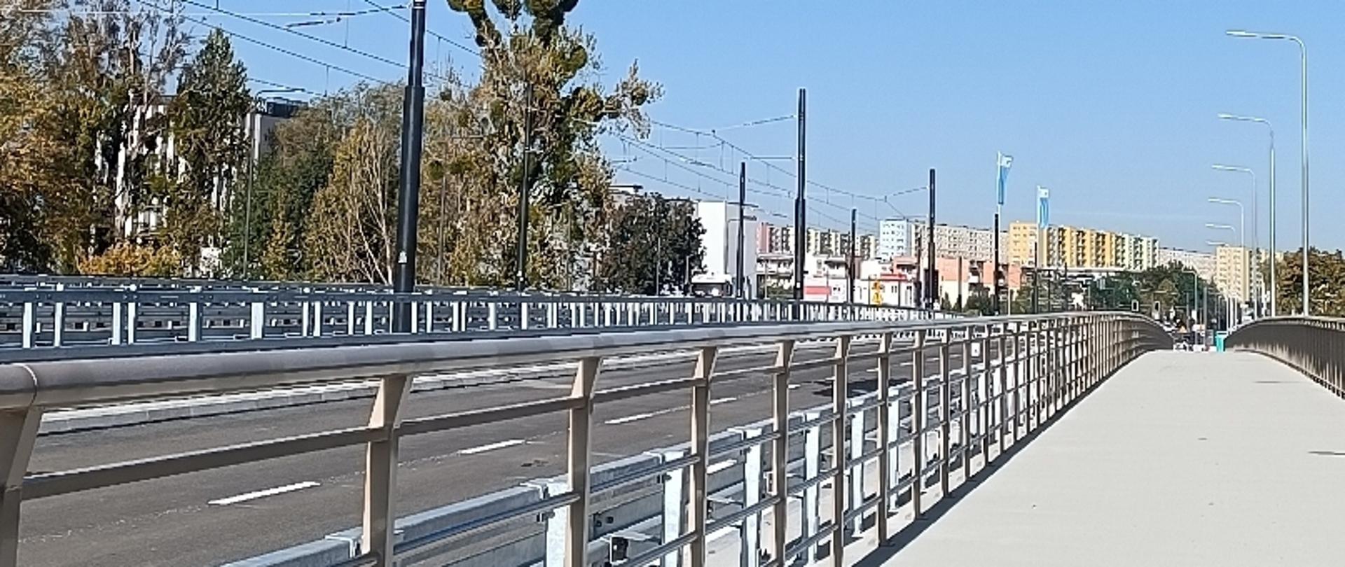 Most z pasami ruchu i chodnikiem, otoczony barierkami i latarniami, z budynkami i drzewami w tle pod błękitnym niebem.