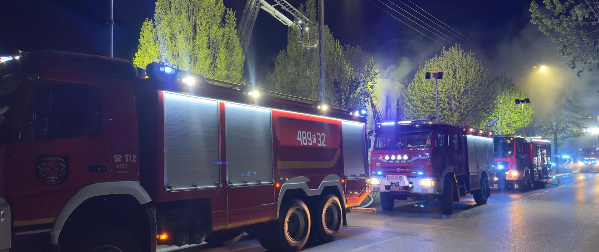Wozy strażackie stoją przed budynkiem. Nad dachem strażacy pracują w koszu podnośnika
