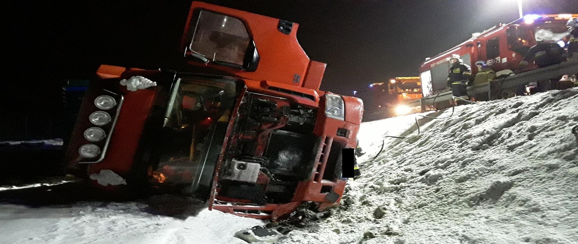 Zdjęcie przedstawia wypadek Tira , który wypadł poza jezdnię autostrady A1 i leży na boku, z prawej stronie widoczny jest samochód pożarniczy i strażacy ratownicy 