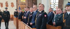 Wnętrze kościoła. W ławkach stoją, komendant główny PSP, komendant Wojewódzki PSP, Minister MSWiA, wojewoda. Przy nich strażacy w mundurach galowych oraz zaproszeni goście.