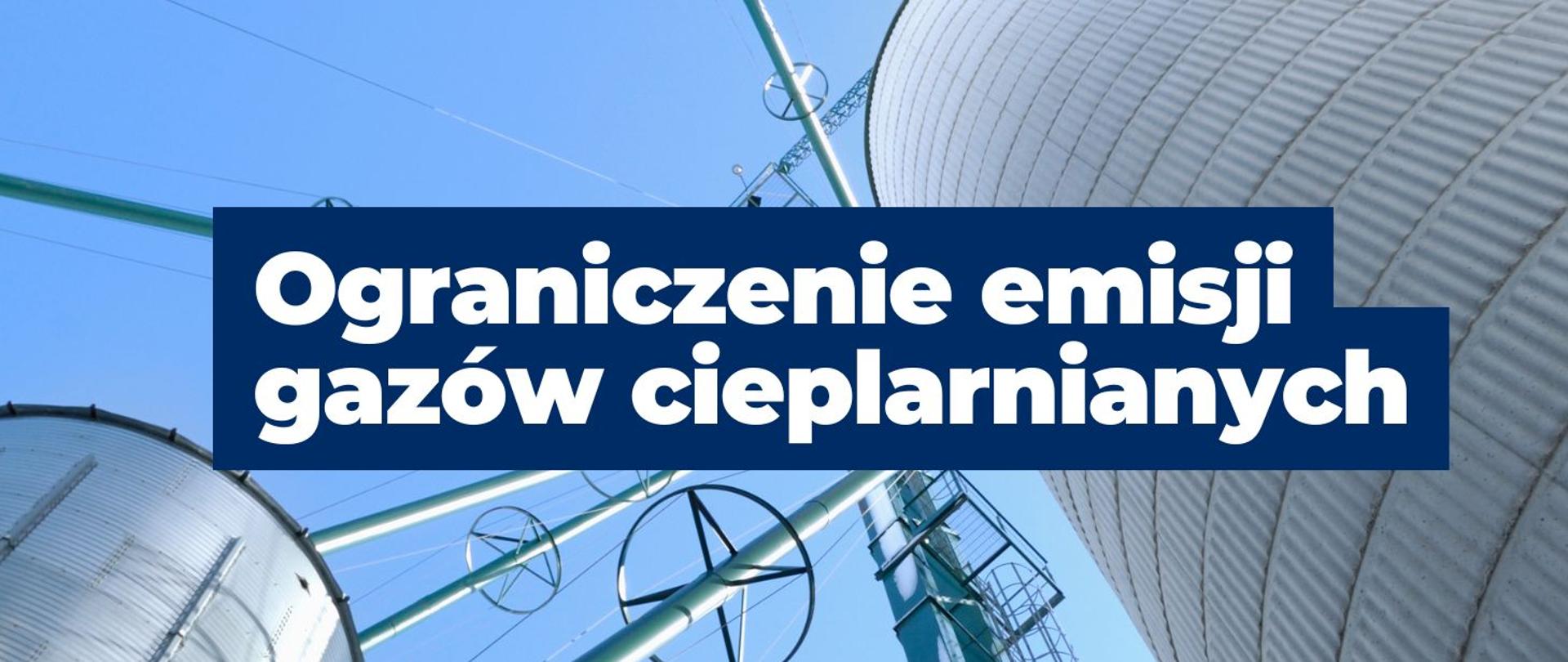 baner z napisem Ograniczenie emisji gazów cieplarnianych . W tle widać zbiorniki przemysłowe. 
