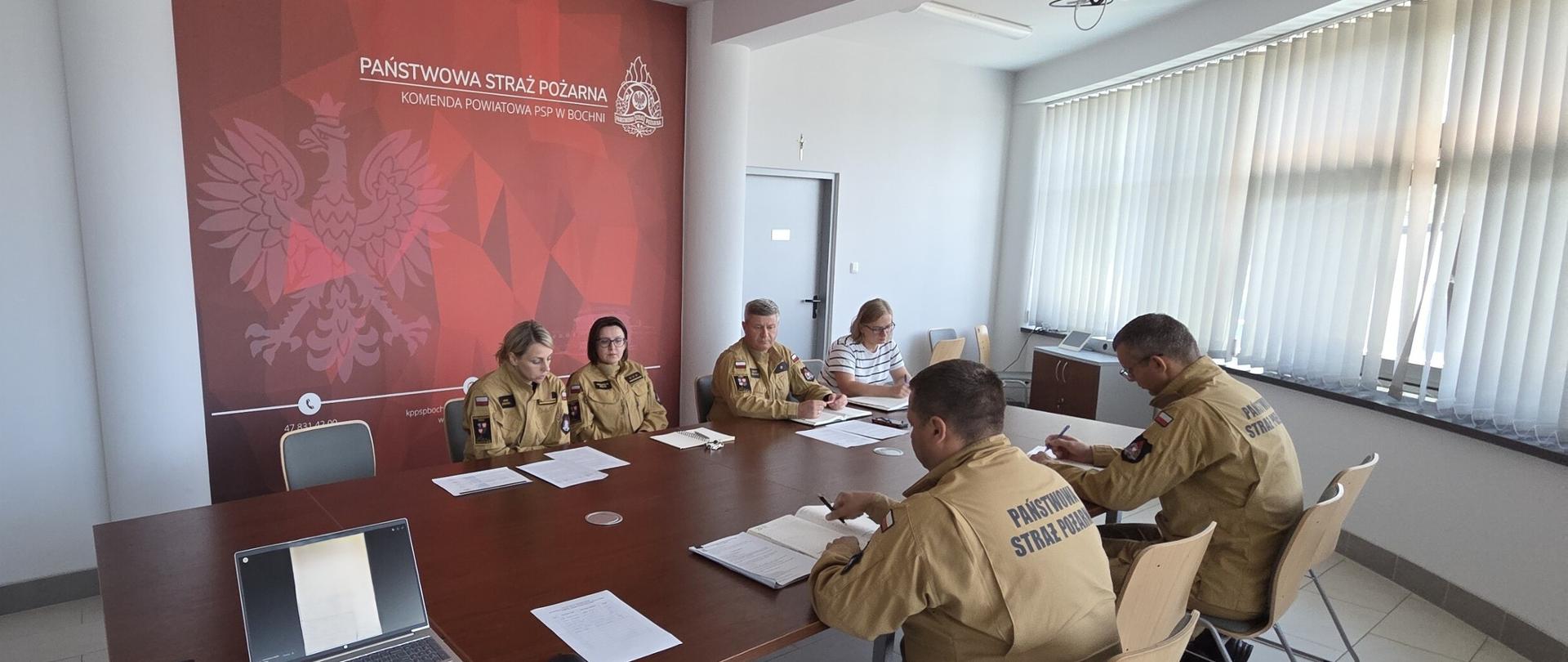 Pięć osób w mundurach oraz jedna osoba w cywilna siedzących wokół konferencyjnego stołu z dokumentami i laptopem, w pokoju z czerwoną ścianą z polskim orłem i napisem Państwowa Straż Pożarna Komenda Powiatowa Państwowej Straży Pożarnej w Bochni.