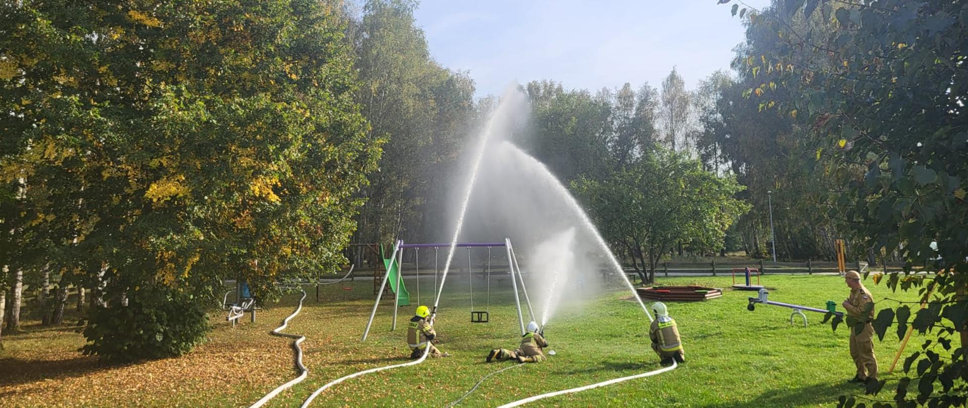 Zdjęcie przedstawia strażaków podczas ćwiczeń z użyciem wody na terenie zielonego parku. Trzech strażaków obsługuje prądy wodne skierowane w górę, tworząc wysokie strumienie i rozpryski. W tle znajduje się plac zabaw, a całość otaczają drzewa w jesiennych barwach. Na trawie widoczne są rozwinięte węże strażackie.