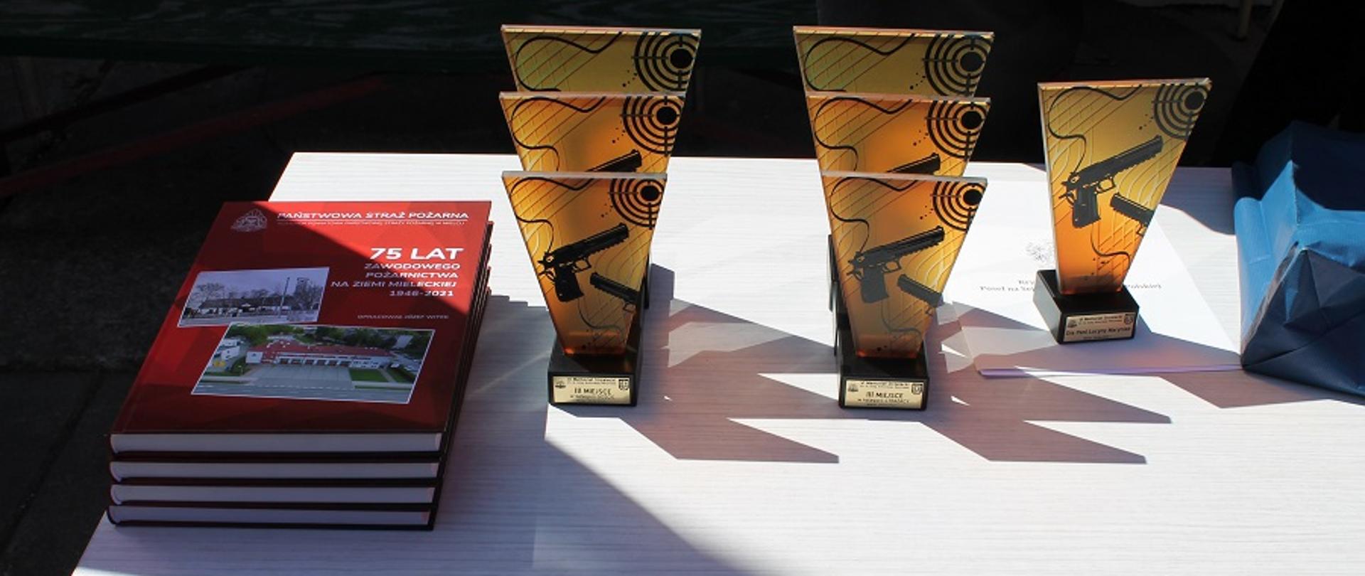Na fotografii biały stolik z pamiątkowymi statuetkami VI Memoriału Strzeleckiego im. st. bryg. Antoniego Maryniaka. Obok leżą albumy wydrukowane z okazji 75 lat zawodowego pożarnictwa na ziemi mieleckiej. 
