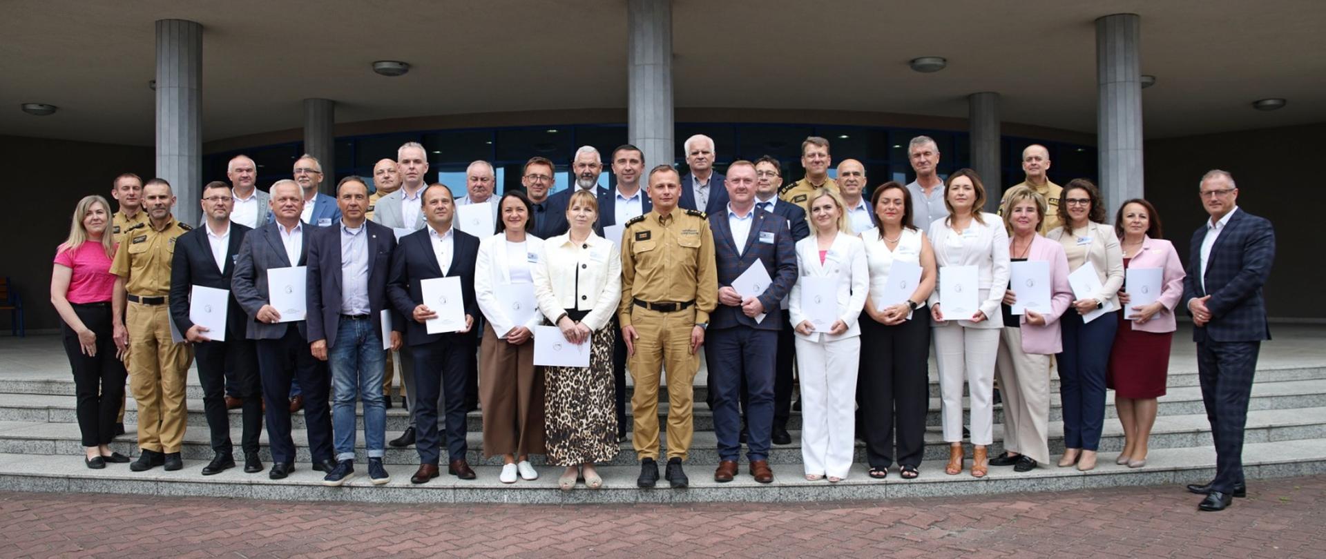 Pamiątkowe zdjęcie grupowe uczestników szkolenia z zakresu ochrony ludności i obrony cywilnej dla osób pełniących funkcję wójtów, burmistrzów i prezydentów miast (23 osoby) w towarzystwie Zastępcy Komendanta Centralnej Szkoły PSP, pięciu oficerów CS PSP, kobiety, przedstawiciela Urzędu Wojewódzkiego z Katowic oraz prelegenta na szkoleniu