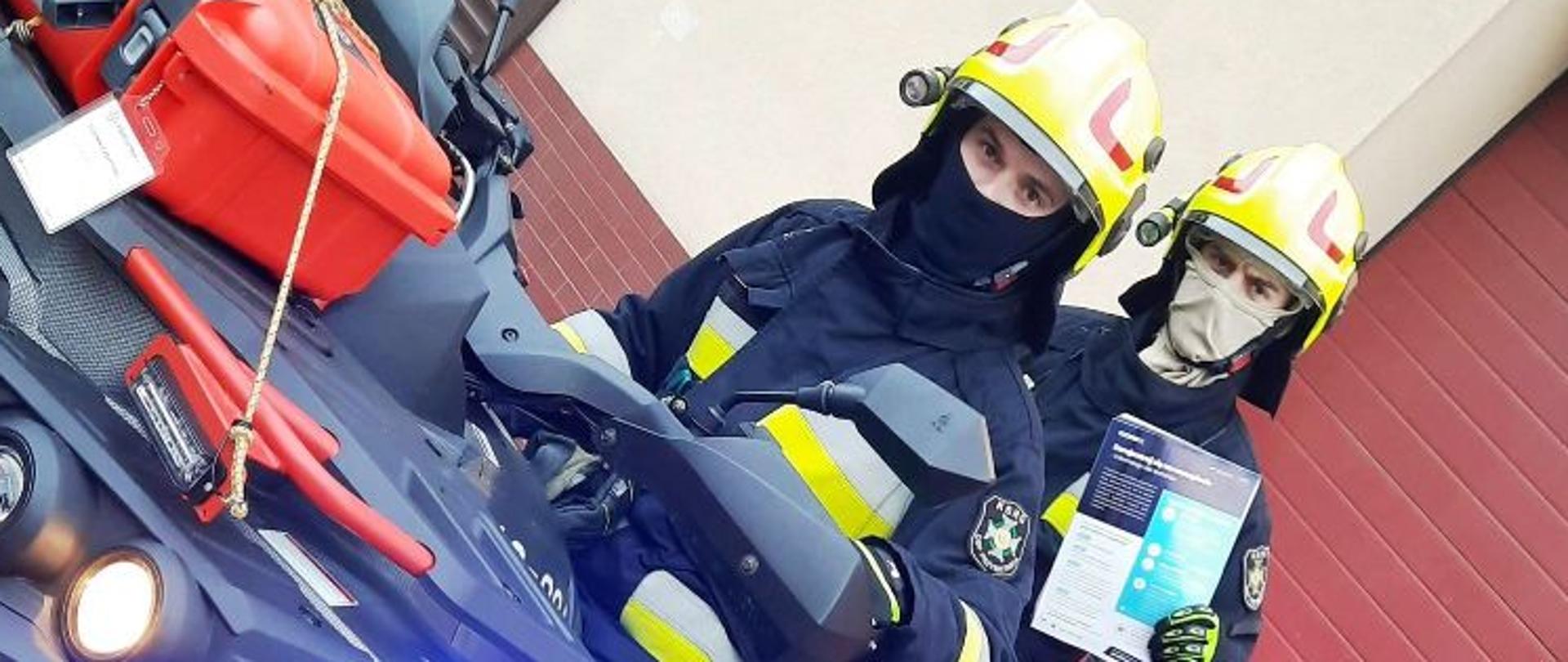 Kolorowa fotografia przedstawia widok pojazdu specjalnego straży pożarnej quda stojącego na utwardzonym placu przed bramą garażową remizy OSP. Na podjeździe siedzą dwie postacie w hełmach, umundurowanych strażaków, którzy w rękach trzymają roznoszone ulotki informacyjne.